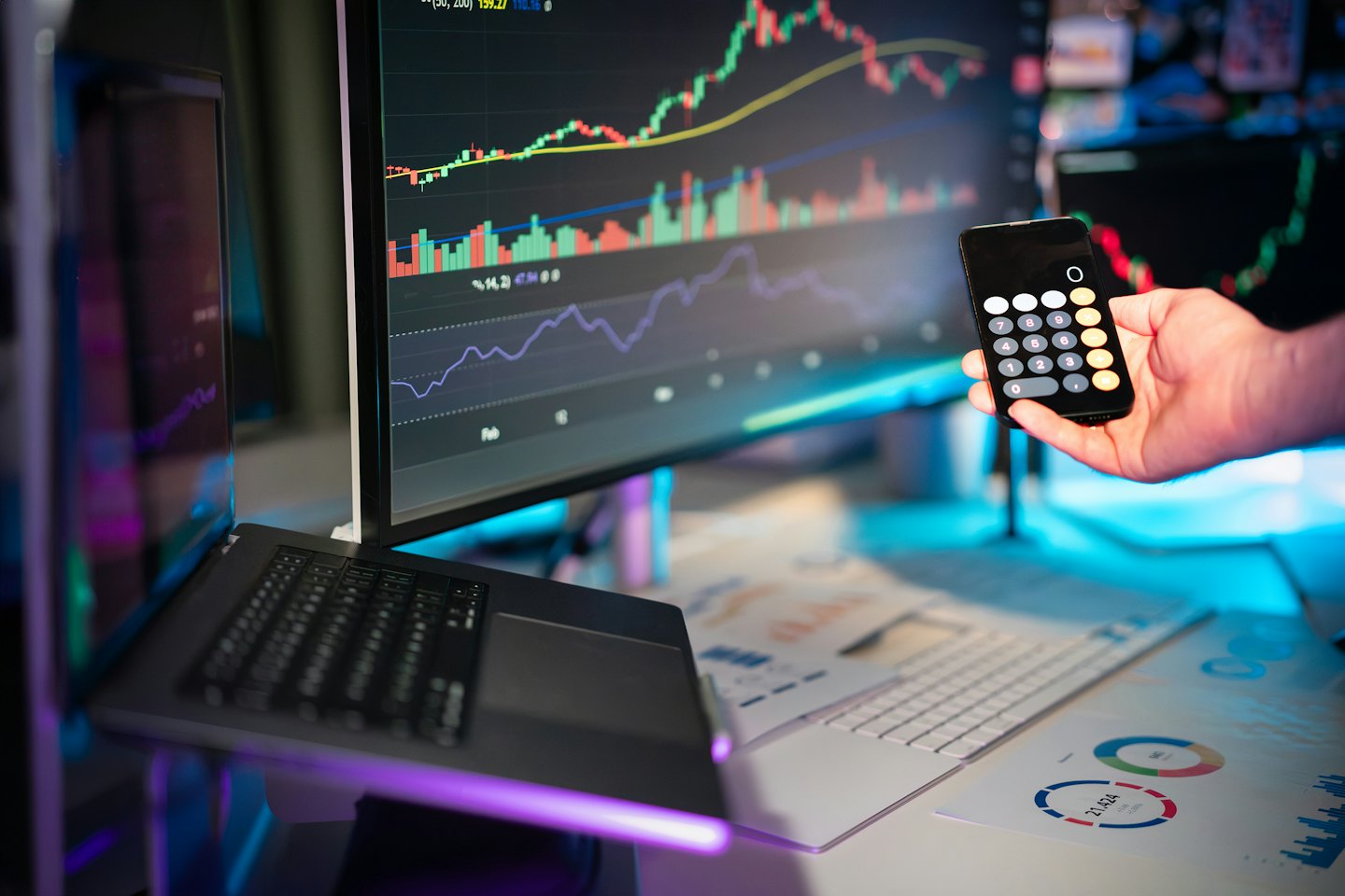 Mano che tiene un telefono con una calcolatrice vicino a dati finanziari su monitor.